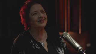 A cantora aparece em primeiro plano ao lado de um microfone. Ao fundo, cortinas de teatro.