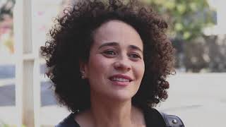 Uma pessoa de cabelos cacheados aparece no centro da imagem. ao fundo, árvores e um poste.