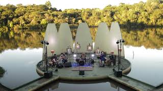 banda aparece em um palco em meio a um lago, com arvores ao fundo