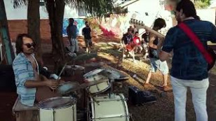 em primeiro plano aparecem dois musicos, um tocando bateria e outro tocando contrabaixo, de costas. ao fundo tem arvores e outras pessoas.