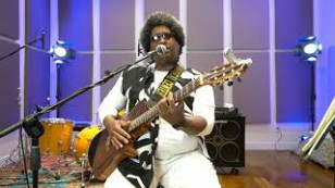 O musico está no centro do plano com uma guitarra e cantando à um microfone. Ao fundo, baterias e caixas de som.