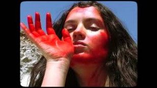 artista ao centro da tela com o ceu ao fundo. a pessoa aparece com as maos e o rosto sujo de tinta