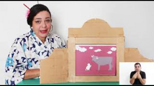 a artista está ao fundo com trajes tipicos japoneses e um cenário de papelão para apresentação em frente