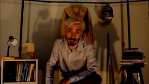 artista no centro do plano vestindo uma camisa social, ao fundo livros e uma luminária