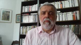 Homem aparece no centro da imagem com uma camisa social. ao fundo, uma estante de livros e ao lado, uma parede com dois quadros.