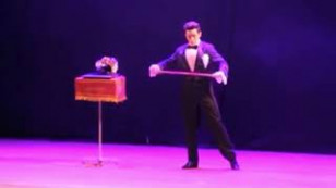 mágico aparece em um palco usando trajes formais, ao lado um objeto não identificado