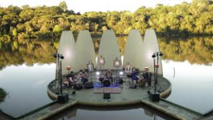 banda aparece em um palco em meio a um lago, com arvores ao fundo