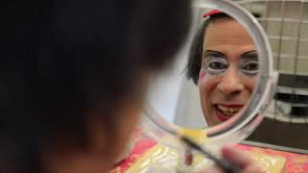 palhaço aparece de costas segurando um espelho que reflete seu rosto sorrindo.