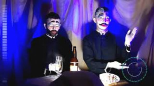 duas pessoas aparecem com maquiagem teatral em frente a um fundo com uma grande cortina, sentadas a uma mesa. na mesa se vê uma garrafa e taças de vidro.