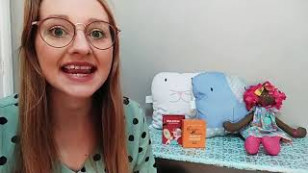 uma mulher aparece do lado esquerdo da tela. no lado direito, uma boneca, duas almofadas e dois livros aparecem.
