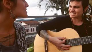 a dupla aparece, com casas ao fundo. um deles toca um violão. 